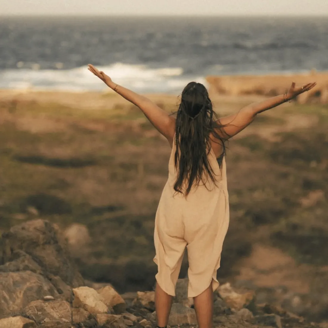 A woman seen from behind with her arms open wide to the sea on a wild, rocky coast
