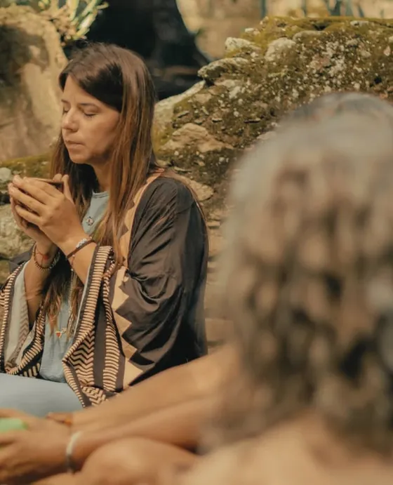 Janine holding a cup of cacao with both hands, eyes closed, sitting in a circle outdoors