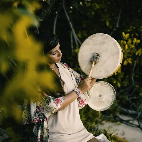 A woman playing a frame drum and a sound instrument among the trees