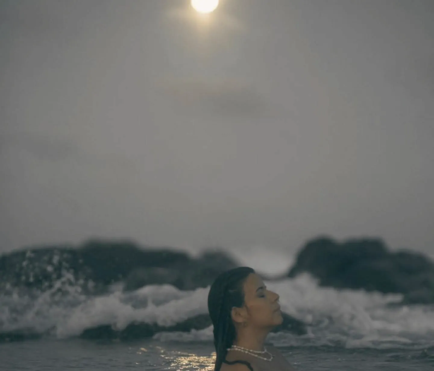A woman in the sea at dusk, eyes closed, waves breaking on the rocks behind her