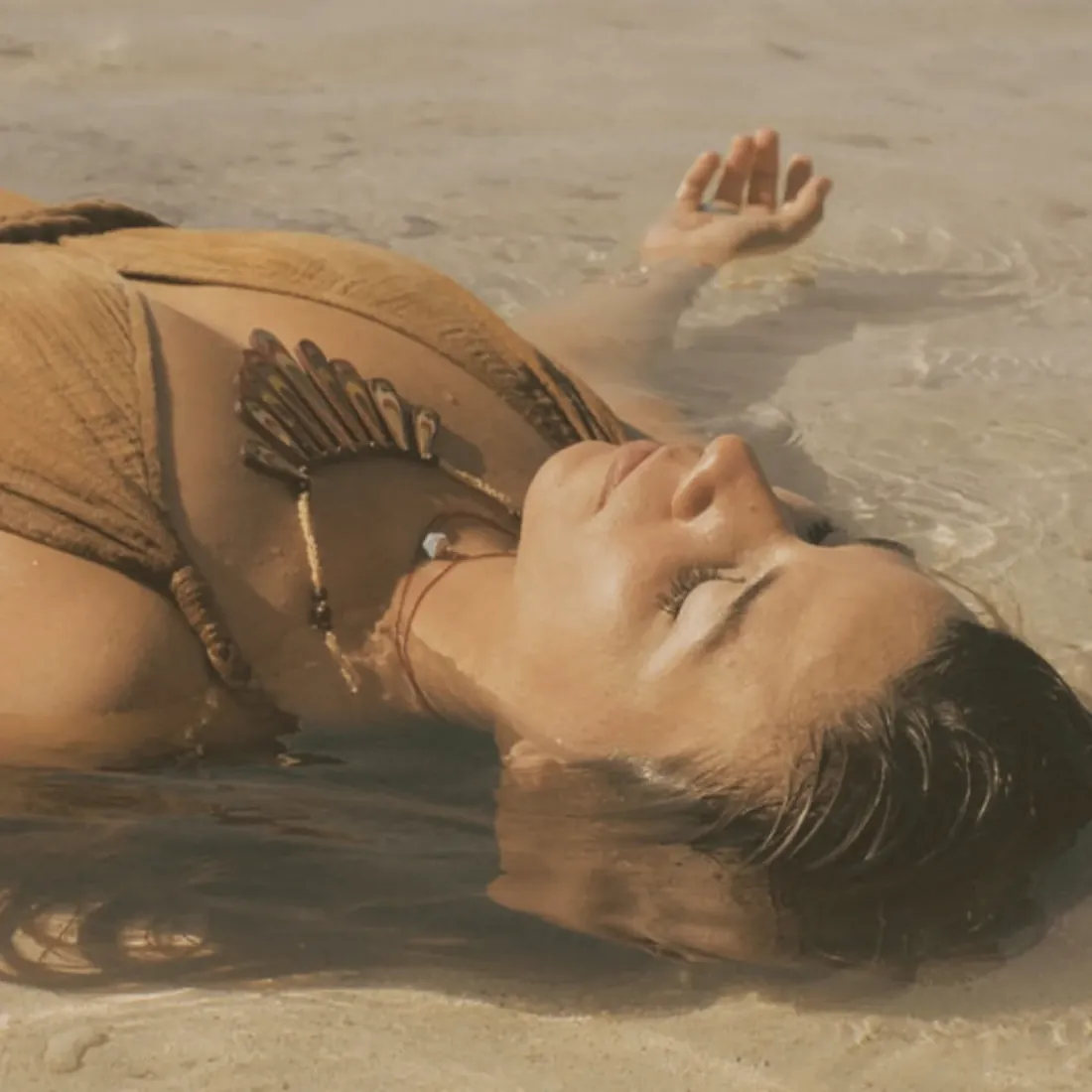 A woman floating on her back in the shallow water of a beach, eyes closed, one hand resting on the surface