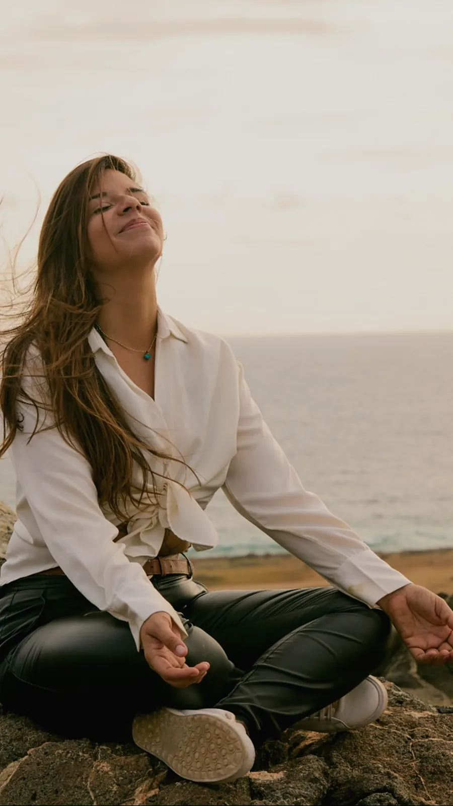 Janine Valecillos meditating on the rocks above the sea in Aruba, eyes closed, smiling