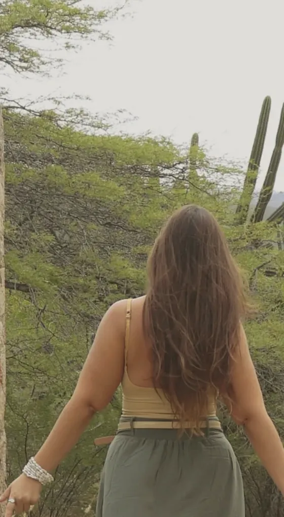 A woman seen from behind, long hair down her back, walking among green shrubs and tall cacti
